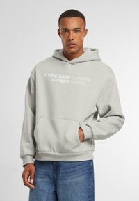 Lichtgrijze hoodie met een voorzak en ribgebreide manchetten. Bevat witte tekst: "APPRECIATE Everything EXPECT Nothing." Zachte stofstructuur.