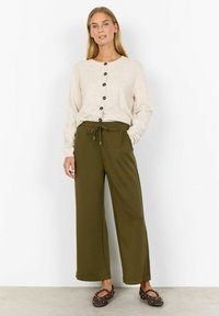 Light beige knitted cardigan with black buttons paired with loose, dark olive-green pants and brown studded flats. Smooth textures and relaxed fit.