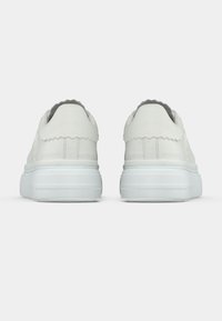 Λευκά αθλητικά παπούτσια με λείο δέρμα φινίρισμα, λεπτομέρεια με κυματοειδή τακούνι και χοντρή λευκή σόλα από καουτσούκ. Μινιμαλιστικός σχεδιασμός χωρίς ορατό λογότυπο.