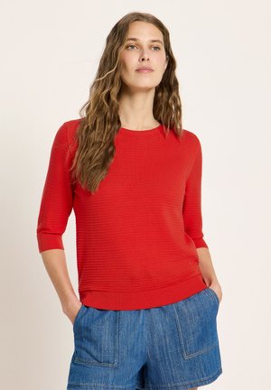 Frau mit langen, welligen Haaren, die ein rotes, strukturiertes Kurzarmtop und blaue Jeansshorts trägt, Hände in den Taschen, neutraler Hintergrund.