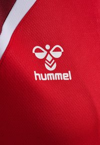 White hummel bee logo and text on red textured fabric, with a white and burgundy stripe on the top left corner.