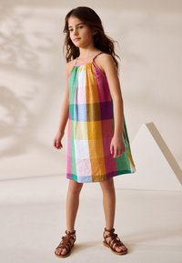 Robe d'été en coton multicolore avec un col froncé, présentant un motif à carreaux en rose, vert, jaune et bleu, assortie à des sandales marron.
