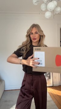 Scatola di cartone marrone con un'etichetta di spedizione, tenuta da una persona con un maglione nero e pantaloni color bordeaux, ambientata in una stanza chiara.