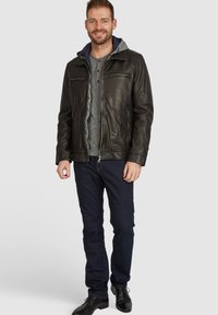Chaqueta de cuero negra con capucha de forro de felpa gris, dos bolsillos laterales y dos bolsillos con cremallera en el pecho, llevada sobre una camiseta henley gris y jeans.