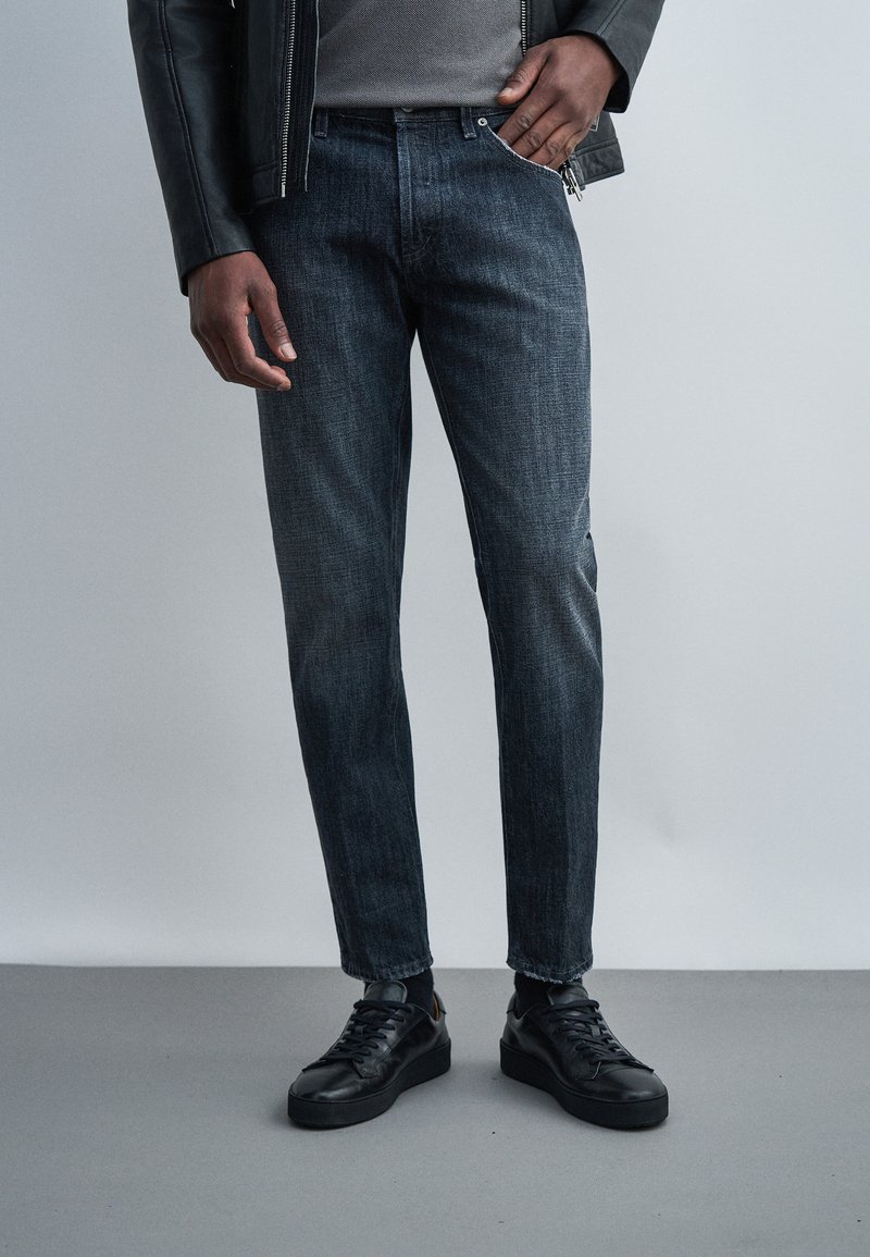 Άνδρας που φοράει σκούρο τζιν, μαύρο δερμάτινο μπουφάν, γκρι πουλόβερ και μαύρα αθλητικά παπούτσια, στέκεται μπροστά σε απλό ανοιχτό φόντο.