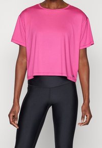 En person i en lös rosafärgad kort t-shirt och tighta svarta högmidjade leggings, stående mot en neutral bakgrund.