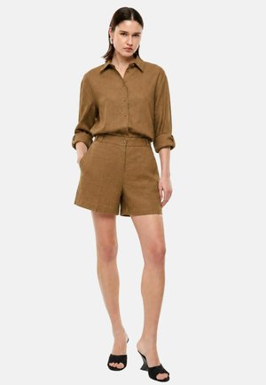 Donna in piedi indossa una camicia marrone abbottonata con maniche arrotolate, pantaloncini marroni coordinati, sandali neri con tacco e orecchini a cerchio d’argento.