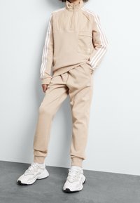 Beige pulovr s čtvrtovým zipem a bílými akcenty ve formě tří pruhů, v kombinaci s odpovídajícími tepláky; zahrnuje přední kapsu a detail loga.