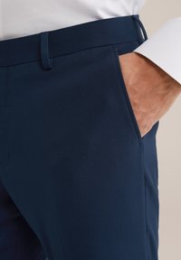 Main d'un homme placée dans la poche avant d'un pantalon formel bleu marine porté avec une chemise blanche.