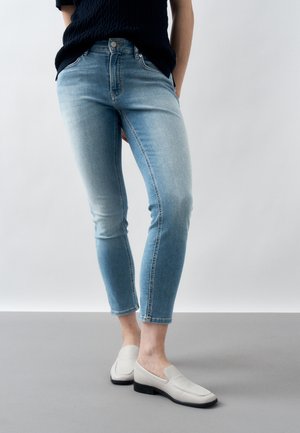 Άτομο που φοράει ανοιχτό μπλε ψηλόμεσο τζιν παντελόνι, μαύρο τοπ και λευκά παπούτσια slip-on, στέκεται σε γκρι πάτωμα.