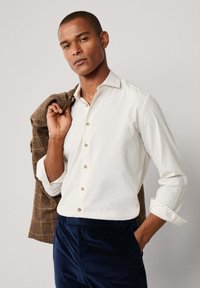 Witte button-up shirt met een zachte textuur, voorzien van een kraag en houten knopen. Gecombineerd met donkerblauwe corduroy broeken en een bruine geblokte blazer.