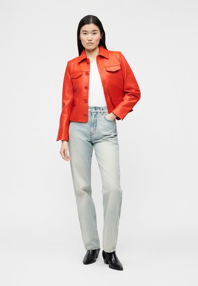 Γυναίκα όρθια με το χέρι στην τσέπη, φορώντας μια φωτεινή κόκκινη δερμάτινη μπουφάν, λευκό τοπ, ανοιχτό μπλε τζιν και μαύρες μποτάκια στο αστράγαλο.