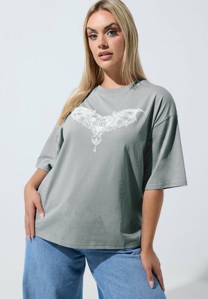 Vrouw met lang blond haar, gekleed in een losse grijze T-shirt met een witte gevleugelde hartgrafiek en lichtblauwe spijkerbroek, staand met één hand op de heup.