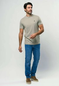 Gai bēšas krāsas īsām piedurknēm T-krekls ar mazu tumšu logo, apvienots ar zilu džinsus un brūnām augstām kedām, stāv uz gaiša fona.