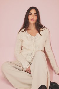 Cárdigan de punto color crema claro con un escote con volantes, cierre de botones y un intrincado detalle de encaje, combinado con pantalones anchos color beige.