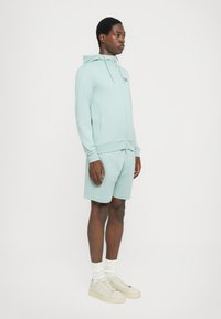 Lys myntegrønn hettegenser med frontglidelås og lommer, sammen med matchende shorts. Skoene er beige med en teksturert såle, og hvite sokker er synlige.