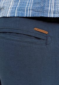 Gros plan sur un pantalon bleu marine avec une poche arrière et une petite étiquette marron de la marque Jack & Jones, associé à une chemise rayée bleue et blanche.