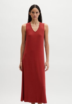 Kobieta o długich ciemnych włosach, ubrana w bezrękawową, czerwonobrązową suknię maxi z dekoltem w serek, stojąca na neutralnym jasnym tle.