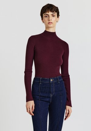 Burgunderfarbener, gerippter Rollkragenpullover mit langen Ärmeln, kombiniert mit hochtaillierten, dunklen Jeans aus Denim mit vorderen Nähten und einem Knopfverschluss.