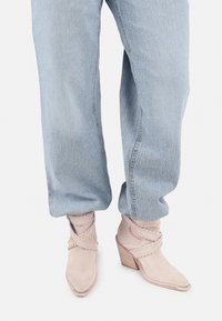 Botines de ante beige con puntera afilada y acentos envolventes, combinados con jeans de mezclilla de corte holgado en azul claro.