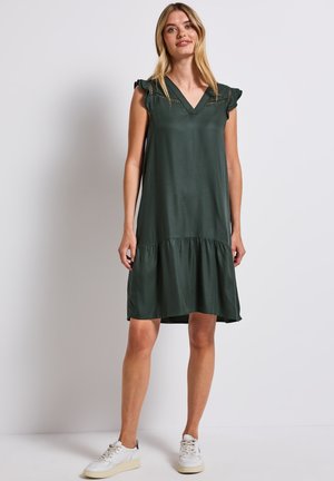 Robe sans manches vert foncé avec un col en V, accents de volants sur les épaules et un ourlet raccourci froncé. Portée avec des baskets blanches.