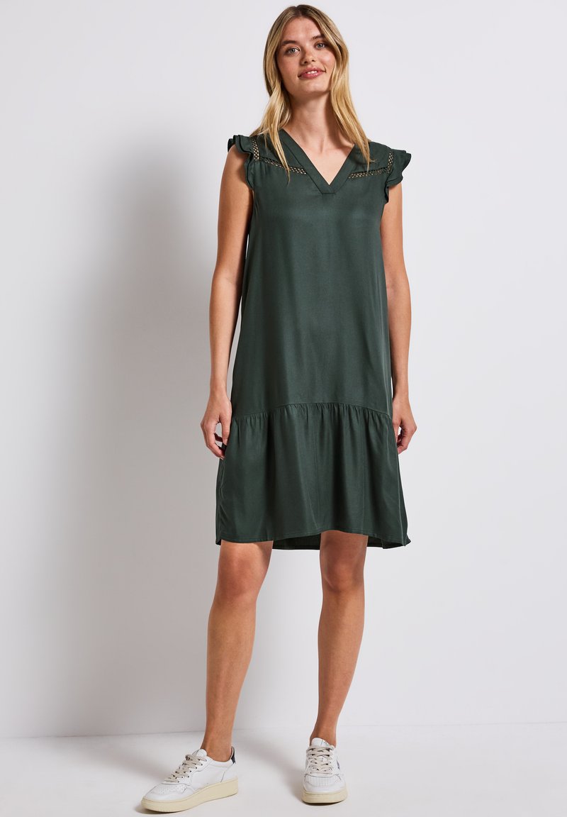 Ciemnozielona sukienka bez rękawów z dekoltem w serek, z falbanami na ramionach oraz marszczonym, warstwowym dołem. Noszona z białymi trampkami.