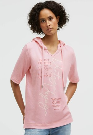 Woman with curly hair wearing a pink short-sleeve hoodie with printed text and light blue jeans, standing against a plain background.