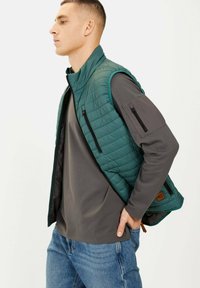 Grüne gesteppte Weste mit hohem Kragen, ausgestattet mit Reißverschlusstaschen und einem grauen Langarmhemd darunter. Eine blaue Jeans rundet das Outfit ab.