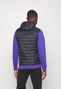 Černá polstrovaná vesta s kapucí, s horizontálním prošíváním, nošená přes fialovou mikinu, s viditelnými černými kalhotami. Minimalistický design.