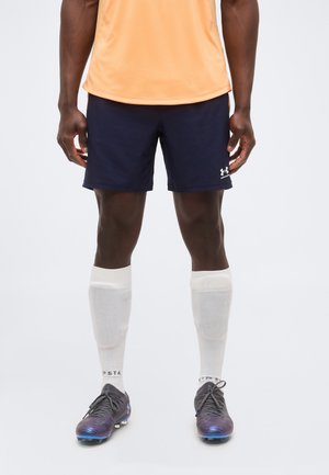 Athlète portant un t-shirt orange, un short Under Armour bleu marine, des chaussettes blanches montantes et des crampons de football noir et violet, debout sur un fond blanc.