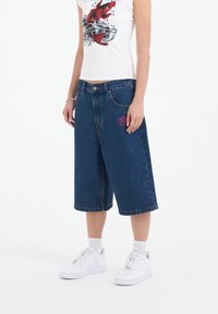 Blue denim culotte shorts with a relaxed fit, featuring front pockets and a pink embroidered detail. Paired with white sneakers.