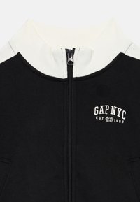 Svart hettejakke med hvit krage, med "GAP NYC"-logo i hvitt, og lommer på hver side. Myk stoff, atletisk design.