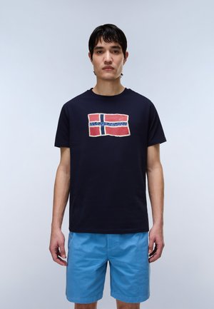 Mlad moški, ki nosi mornarsko modro majico s rdeče-belim motivom zastave in modre kratke hlače, stoji pred enostavnim svetlim ozadjem.