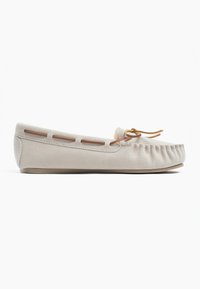 Mocasin de ante beige con puntera redondeada, costuras decorativas y un lazo delgado de cuero en la parte delantera. Presenta detalles laterales recortados.