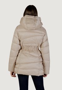 Giacca a vento beige con design trapuntato, vita regolabile con coulisse e cappuccio; presenta maniche lunghe e una texture liscia e lucida.