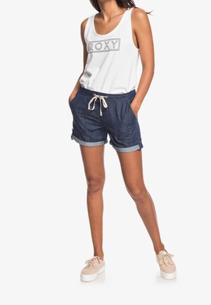 Vrouw die een witte mouwloze top draagt met het "ROXY"-logo, marineblauwe opgerolde korte broek met trekkoord en beige sneakers, staand met handen in de zakken.