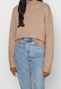 Pull crème court avec un col montant, des poignets côtelés et une texture douce. Associé à un jean en denim bleu clair taille haute.