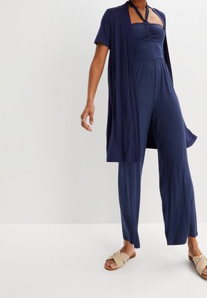 Mørkeblå jumpsuit med halterneck, rynket bodice og vidde ben, parret med en matchende kortærmet cardigan og beige sandaler.