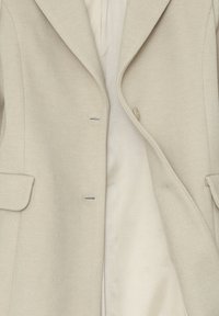 Cappotto in lana beige con texture, colletto a riviera, tasche a pattina e una fodera interna di colore crema, parzialmente aperto che mostra i dettagli del tessuto.