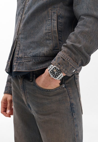 Tume teksajope peene tekstuuriga, sobitatud tume teksapükstega. Käel on hõbedane kell musta sihverplaadiga, millel on geomeetriline disain.