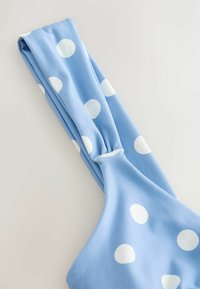 Tissu bleu clair à pois blancs rassemblé et torsadé, probablement partie d'un vêtement ou accessoire sur un fond neutre.