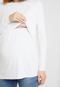 Blusa de manga comprida branca com uma abertura para acesso à amamentação, feita de tecido suave; combinada com jeans azuis. Textura suave, corte relaxado.