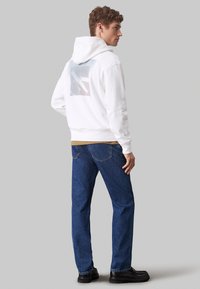 Junger Mann in weißem Hoodie mit Grafik auf dem Rücken, blauen Jeans und schwarzen Schuhen, der steht und nach rechts auf einem schlichten grauen Hintergrund schaut.