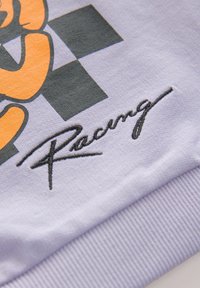 Lys lilla stof har et orange tegneserie-grafik og sort ternet mønster, med broderet "Racing" i kursiv på den nederste kant.