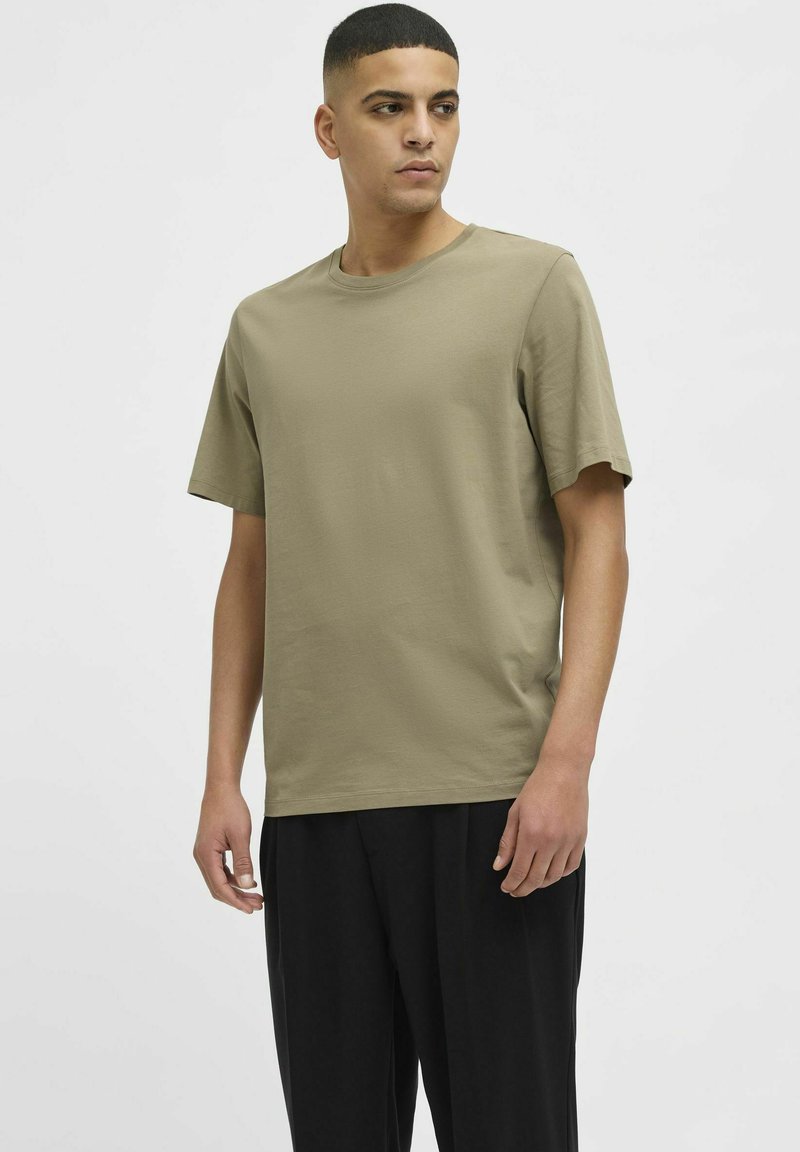 Camiseta de manga corta en color verde oliva, fabricada en suave algodón, con un clásico cuello redondo y un corte relajado. Combinada con pantalones negros.