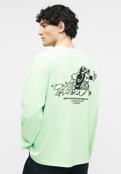 Άνδρας με σγουρά μαλλιά που φοράει ανοιχτοπράσινη μπλούζα με μαύρο σχέδιο ενός τρέχοντος χαρακτήρα και τον αριθμό 26 στην πλάτη.