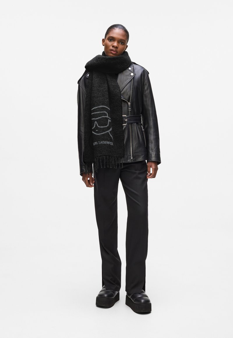 Zwarte leren jas met een cinched taille, gecombineerd met een donkere sjaal met een lichtgrijze graphic. Model draagt zwarte wijde broeken en plateau schoenen.