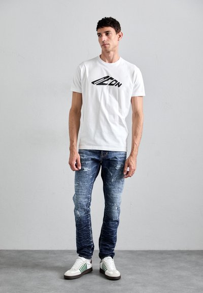 White cotton t-shirt with a black graphic logo on the front, paired with dark blue distressed jeans and white sneakers with green accents.