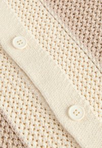 Tissu tricoté texturé avec une bande crème boutonnée comportant deux boutons blancs ronds sur un matériau tissé beige et crème.