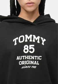 Čierna mikina s kapucňou s potlačou na prednej strane. Text: "TOMMY 85 AUTHENTIC ORIGINAL since 1985." Má kapucňu so sťahovacou šnúrkou a mäkkú textúru látky.
